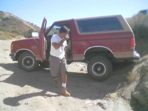 1990 Ford Bronco - man in a white shirt and shorts steps out of the open door of a red two-tone Ford Bronco parked in a sandy desert wash.