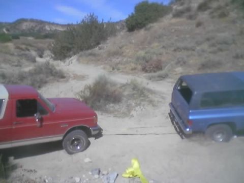 1990 Ford Bronco - red Ford truck is connected by a tow strap to a blue SUV on a sandy desert trail, with a yellow marker on the ground between them.