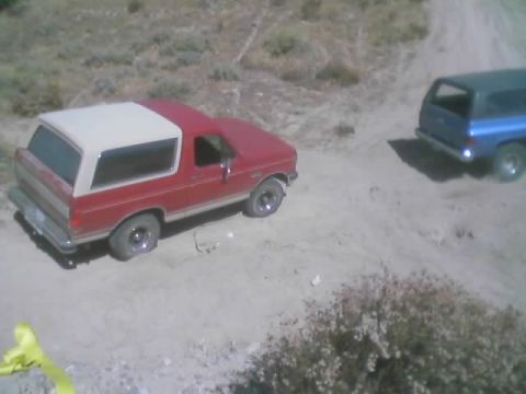 1990 Ford Bronco - red-and-white two-door SUV sits on a sandy off-road trail beside a blue SUV among desert scrub.