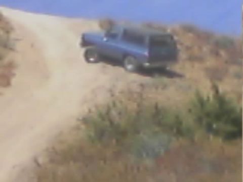1990 Ford Bronco - blue two-door SUV travels along a dirt ridgeline trail surrounded by dry brush under a clear sky.