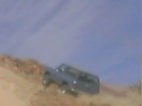 1990 Ford Bronco - dark-colored SUV climbs a steep dirt embankment in a blurry distant shot under a blue sky.