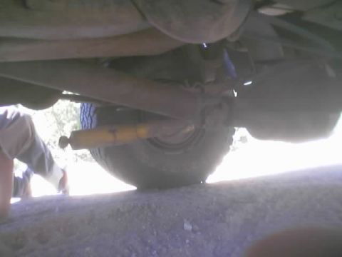 1990 Ford Bronco - low ground-level view beneath a vehicle showing the axle, tire, and a yellow shock absorber.