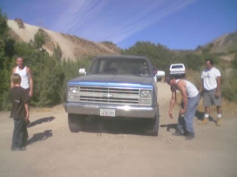 1990 Ford Bronco - blue-and-silver square-body Chevrolet pickup sits on a dirt road as several people stand around it.