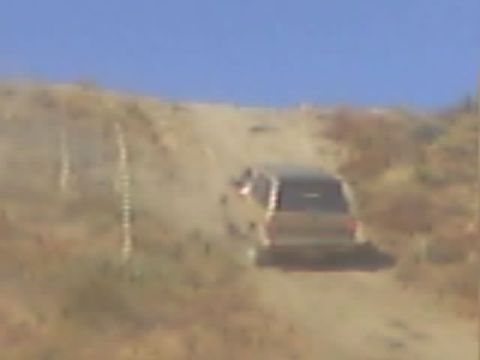 1990 Ford Bronco - blurry rear view of an SUV climbing a steep, dusty hill trail amid dry vegetation.