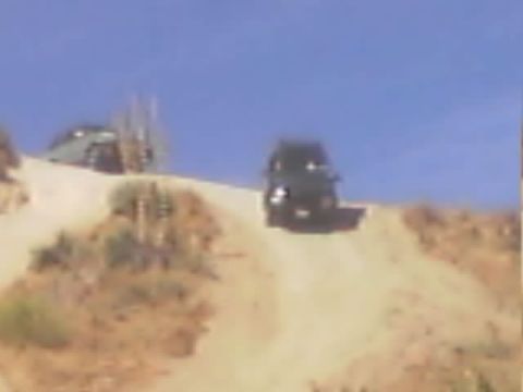 1990 Ford Bronco - Two SUVs sit on a sandy ridge trail under a clear blue sky, one dark vehicle facing the camera.