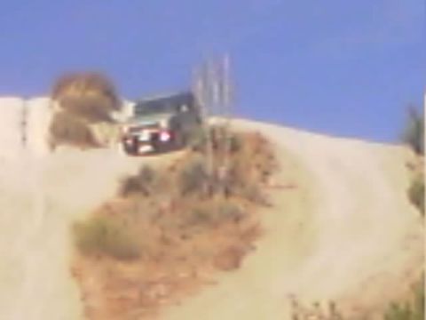 1990 Ford Bronco - dark SUV with its headlights on sits atop a steep sandy hill trail among dry brush.