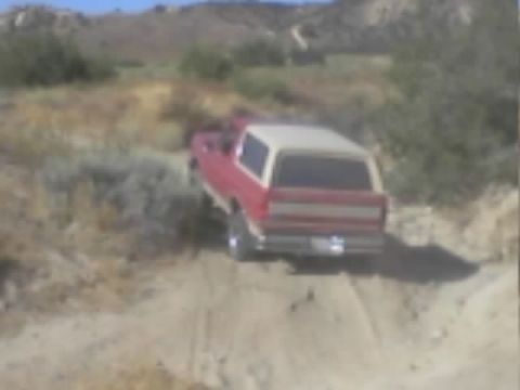 1990 Ford Bronco - red SUV with a white roof climbs a rutted dirt hill amid desert scrub with mountains behind.