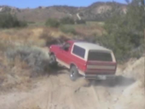 1990 Ford Bronco - red SUV with a white roof climbs a desert dirt trail viewed from behind, with hills in the distance.