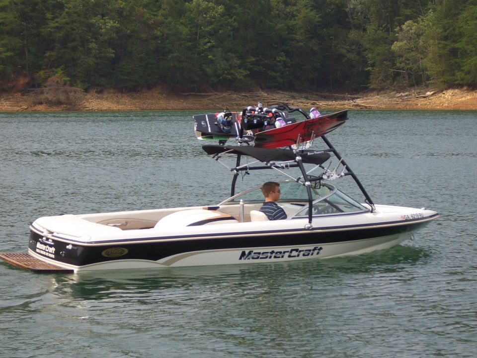 White MasterCraft wakeboard boat with tower and boards on a calm lake, side view, driver aboard.