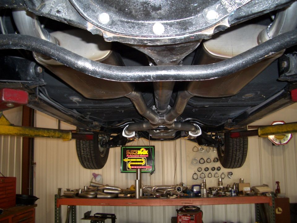 Undercarriage view of a car on a lift, showing dual exhaust system and rear differential. Dyno-Max sign visible