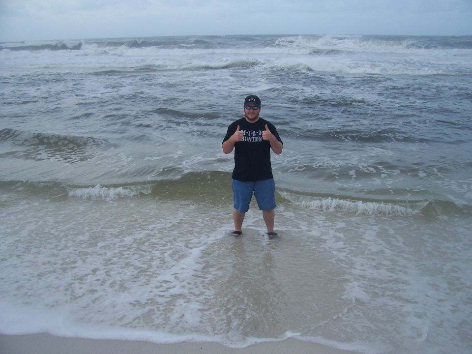 Man in black t-shirt and blue shorts gives two thumbs up while standing in ocean waves