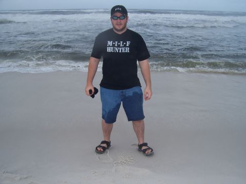 1995 Ford Bronco - A man stands on a sandy beach with waves crashing in the background, wearing a t-shirt with text and denim shorts.