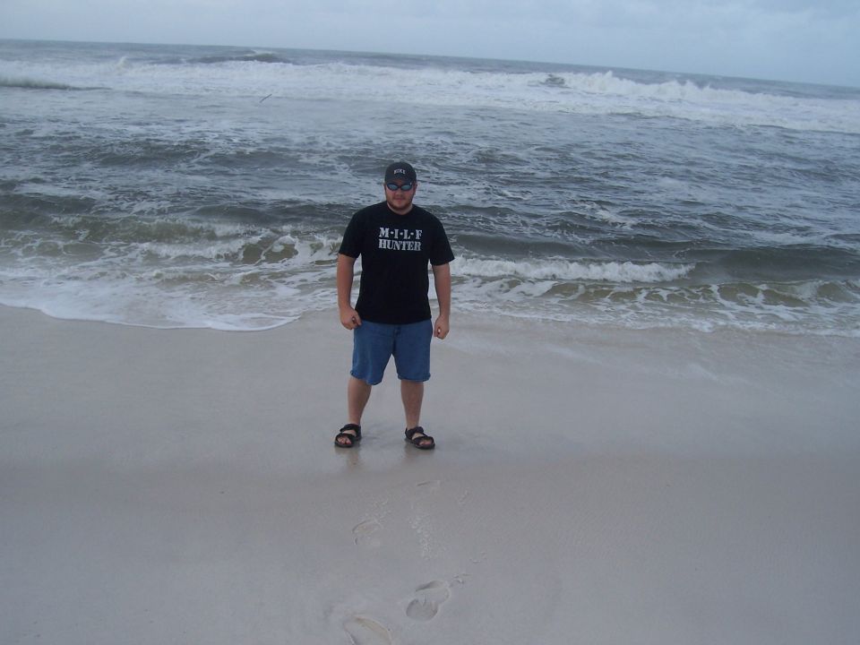 Man wearing a black t-shirt with text and blue shorts stands on a sandy beach with waves in the background