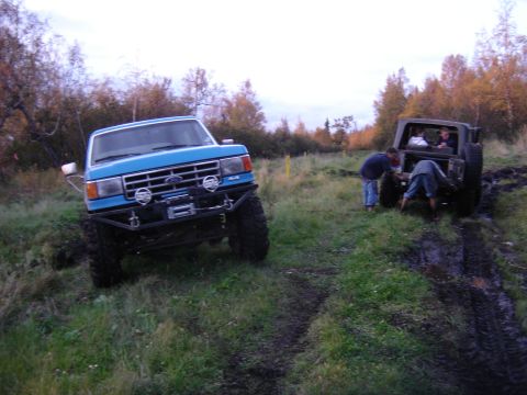 1988 Ford Bronco - Blue lifted Ford truck with a tube front bumper, winch, KC-style driving lights and mud tires sitting on a grassy trail rutted with mud, sh…