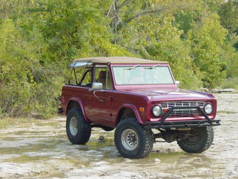 1977 Ford Bronco - This maroon Ford Bronco is equipped with a brush guard and a soft top, seen here in a watery, natural setting.