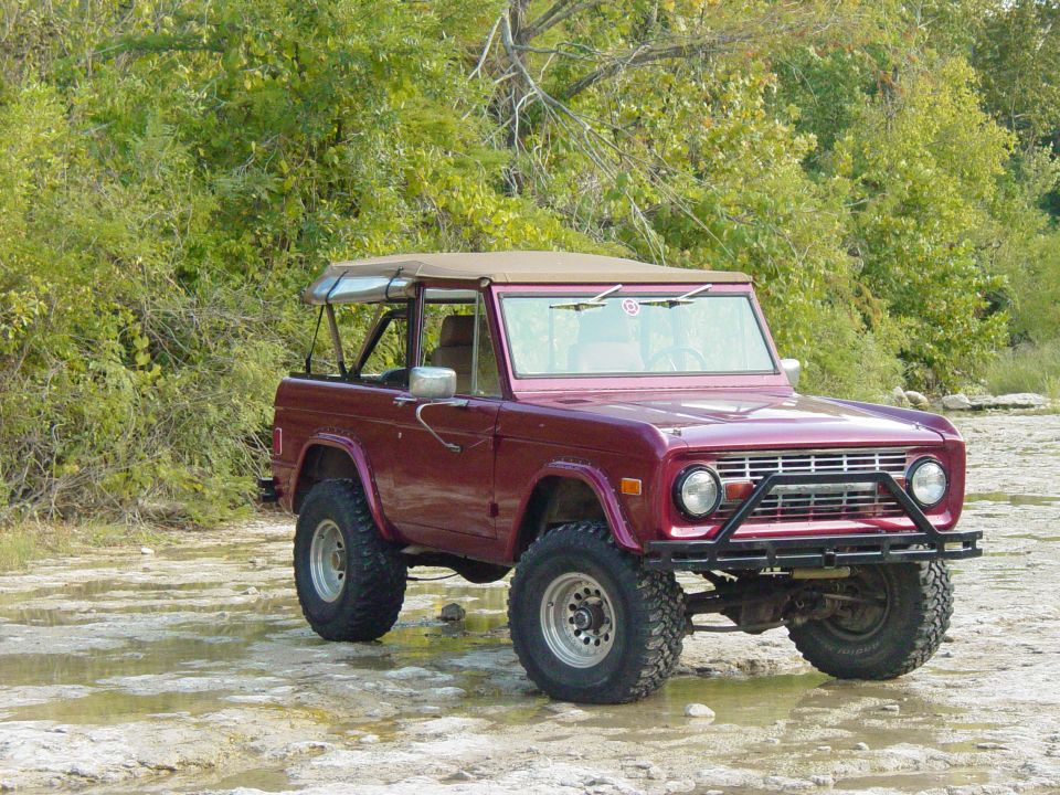 Maroon early Ford Bronco with tan soft top lifted on mud tires