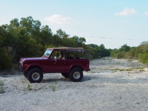 1977 Ford Bronco - This maroon 1977 Ford Bronco, equipped with a tan soft top, is parked in a dry, rocky riverbed surrounded by trees.