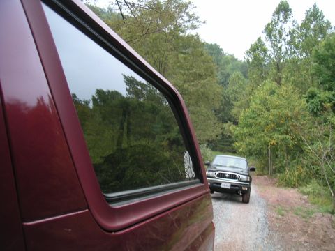 1994 Ford Bronco - A dark SUV is seen on a gravel road, with a maroon SUV in the foreground, surrounded by trees.