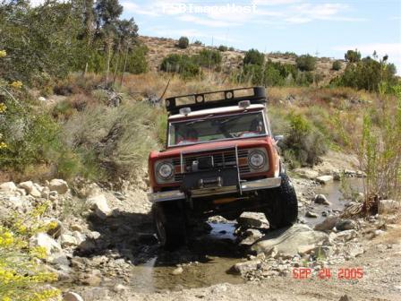 1988 Ford Bronco - A red off-road vehicle drives through a rocky stream on a sunny day.