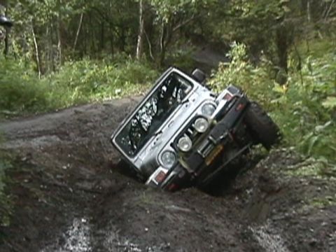 2003 Jeep TJ - An off-road vehicle is stuck at an angle in a muddy rut on a dirt path surrounded by trees.