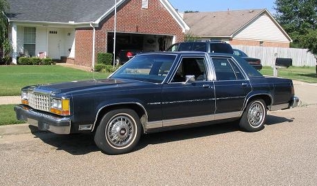 1985 Ford Crown Victoria LTD - This dark blue sedan is parked on a residential street in front of a brick house.