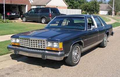 1985 Ford Crown Victoria LTD - A dark blue sedan is parked on the street in front of a house with a garage and a minivan.