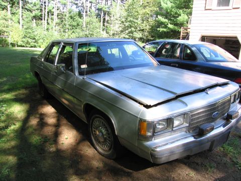 1991 Ford LTD Crown Victoria - A silver Ford Crown Victoria sits in a driveway with its hood ajar, next to a dark-colored sedan.
