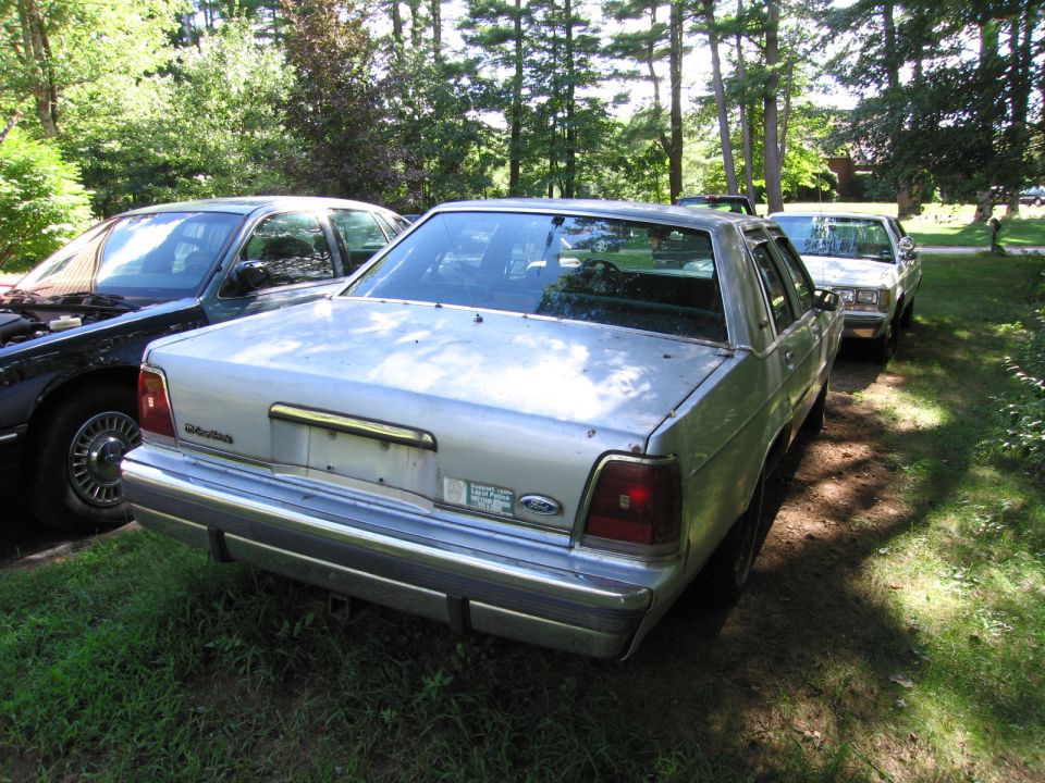 Silver 1990 Ford Crown Victoria sedan, rear 3/4 view, parked in grassy area with trees