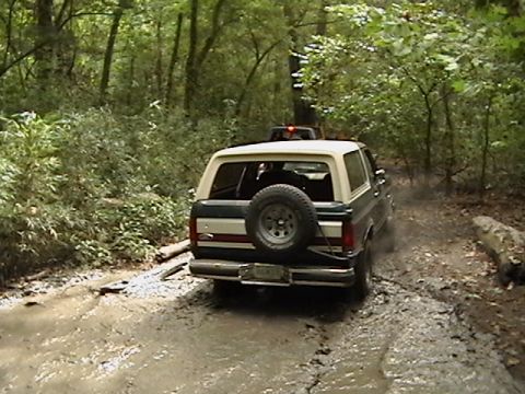 1986 Ford Bronco - A Ford Bronco navigates a muddy trail in a wooded area, with another vehicle following behind.