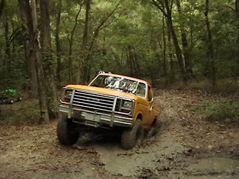 1986 Ford Bronco - This truck is navigating a muddy trail in a forest, with another vehicle partially visible in the background.