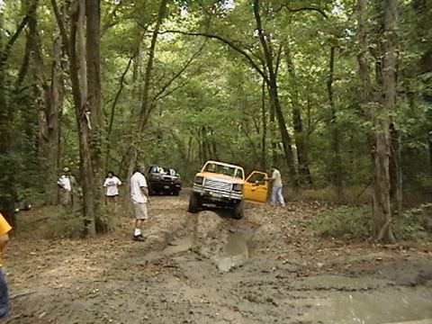 1986 Ford Bronco - A yellow off-road truck navigates a challenging muddy trail with a black truck visible in the background.