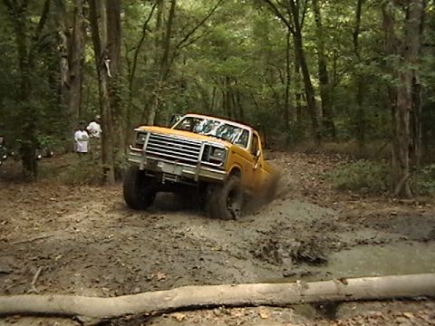 1986 Ford Bronco - A yellow pickup truck navigates a muddy trail in a wooded area.