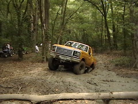 1986 Ford Bronco - A yellow off-road truck navigates a muddy trail, with other vehicles and people visible in the background.