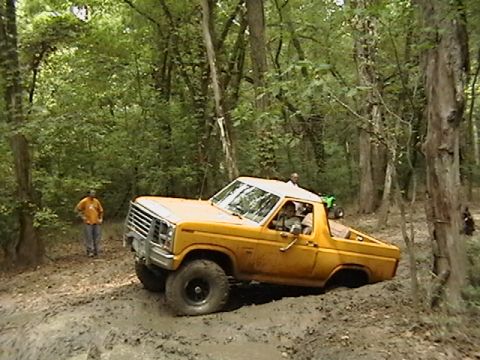 1986 Ford Bronco - Off-road SUV navigating a challenging muddy terrain surrounded by dense forest.