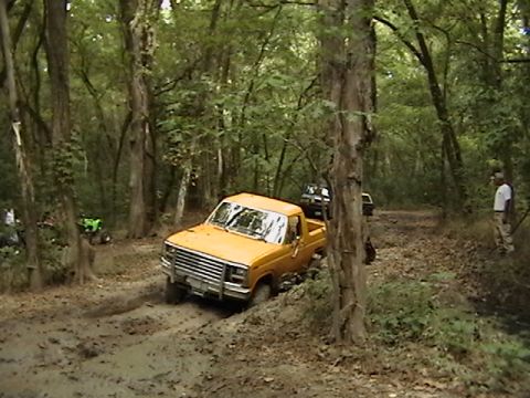 1986 Ford Bronco - A yellow pickup truck navigates a muddy path in a wooded area, with other vehicles nearby.