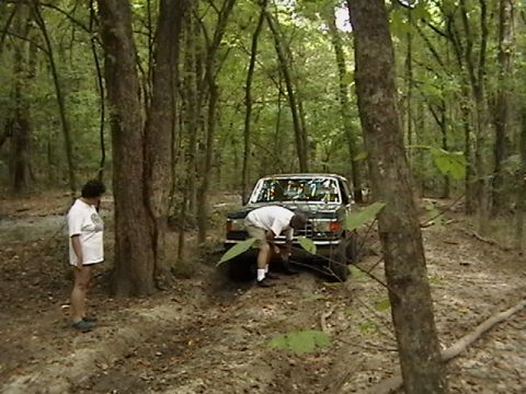 1986 Ford Bronco - A dark-colored Ford Bronco appears to be stuck in the mud while two people attend to it in a wooded area.