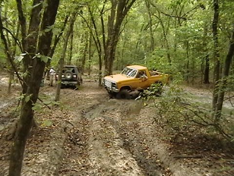 1986 Ford Bronco - Two off-road vehicles, a yellow pickup and a black SUV, are stuck in mud on a wooded trail.