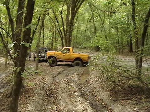 1986 Ford Bronco - Two off-road vehicles navigate a muddy trail through a dense forest.