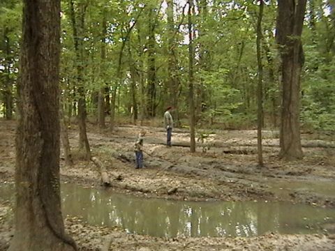 1986 Ford Bronco - A wooded scene with muddy terrain and a small body of water, two individuals in the distance.