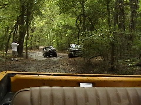 1986 Ford Bronco - Two SUVs navigate a muddy path through a dense forest, with a bystander observing the off-road excursion.