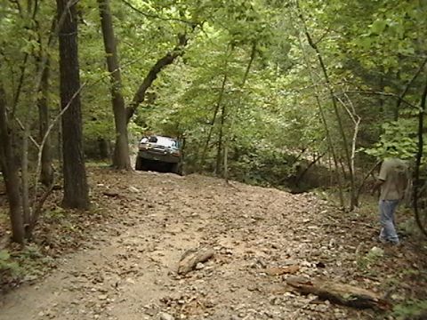 1986 Ford Bronco - An off-road vehicle navigates a challenging dirt path surrounded by dense trees.