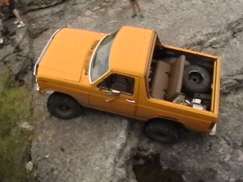 1986 Ford Bronco - This orange SUV is navigating a rocky, off-road environment. A spare tire is visible in the cargo area.