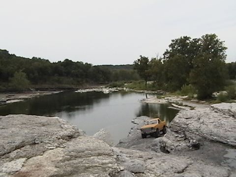 1986 Ford Bronco - A yellow pickup truck is parked on a rocky shore next to a calm body of water.