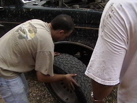 1986 Ford Bronco - A man works on the tire of a dark green SUV, with another person standing nearby.