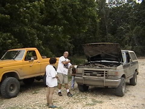 1986 Ford Bronco - Two vehicles are parked on a dirt road, with people standing around the front of a tan Ford Bronco with its hood open.
