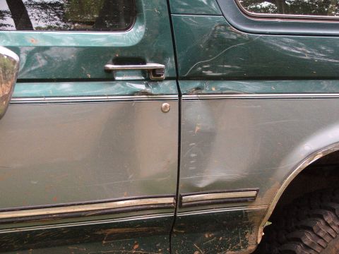 1996 Ford Bronco - Side view of a dented and scratched green and silver SUV, showing the door and part of a tire.