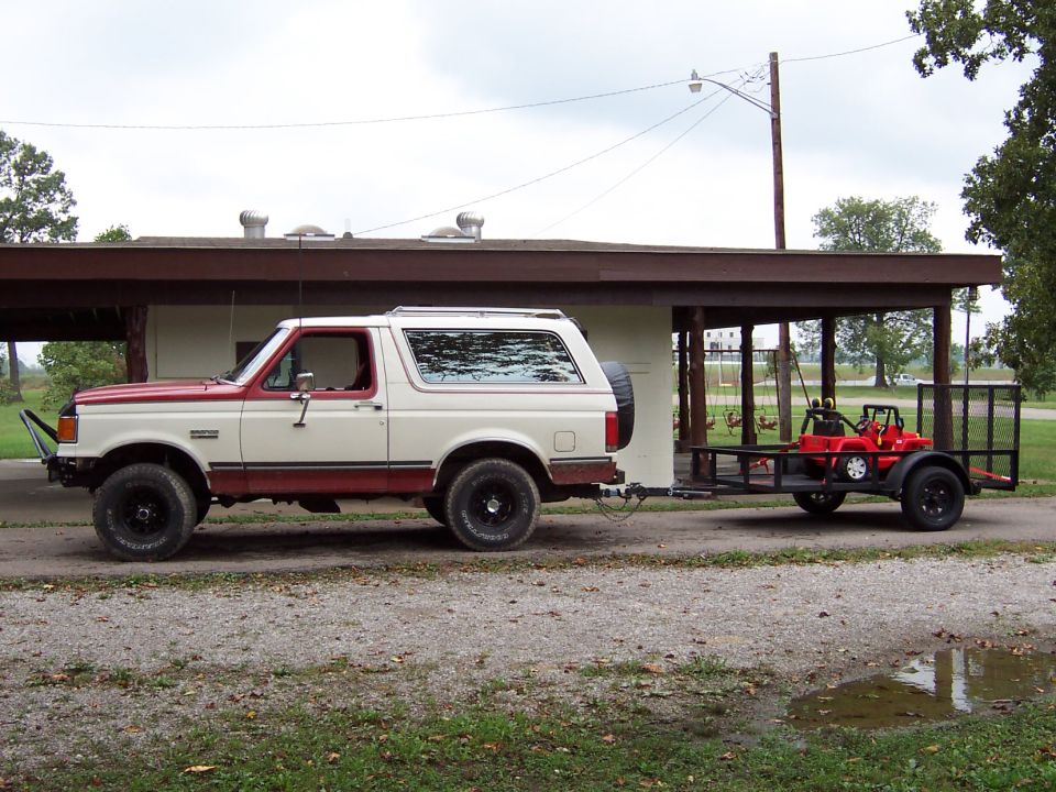 Two-tone Ford Bronco, likely late 80s, towing a trailer with a red Power Wheels car on it
