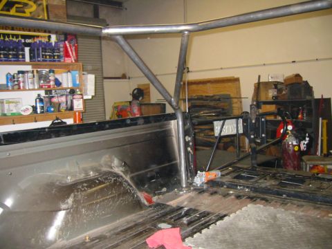 1992 Ford Bronco - Bare-metal roll cage tubing being fitted through the bed of a stripped truck in a home garage, with a rear hoop and diagonal brace passing…