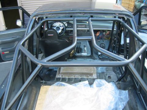 1992 Ford Bronco - View down into the stripped cabin of a Ford truck/Bronco from behind, showing a black-painted multi-point roll cage with door bars, harness…