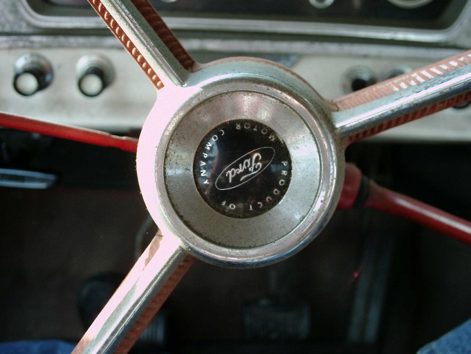 Four-spoke steering wheel hub with chrome horn ring and Ford Motor Company emblem, red rim, dashboard behind